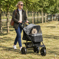 Charger l'image dans la visionneuse de la galerie, A black and gray pet stroller with a small fluffy white dog sitting inside, parked on a grassy lawn next to a body of water with trees in the background.